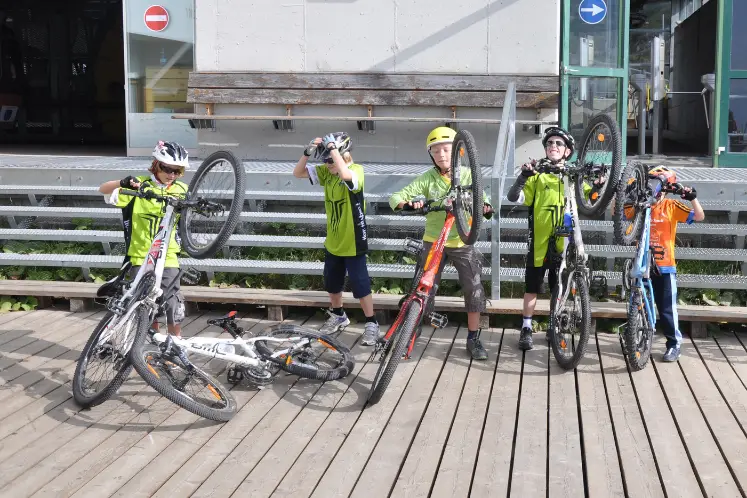 Gruppe von Kindern mit ihren Mountainbikes auf die Hinterräder gestellt steht nebeneinander stolz vor einer Gondelstation. Alle tragen alle Helme und bunte Fahrrad Trikots. Sie haben Spass, alle Kinder haben ein passendes Rad.