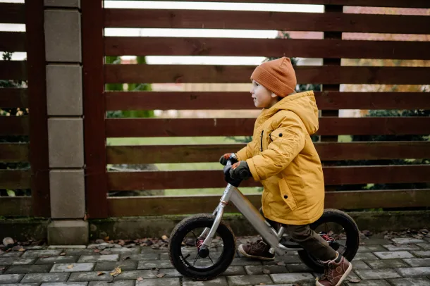 Ein Kind fährt mit seinem Laufrad im Herbst mit Jacke und Mütze an einem Zaun entlang.